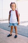Écolier avec sac à dos debout sur la chaussée regardant la caméra en plein soleil — Photo de stock