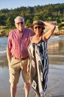 Sonriente pareja de ancianos descalzos en gafas de sol de pie en la playa de arena húmeda y disfrutando de un día soleado - foto de stock
