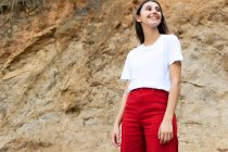 Young contemplative happy female adolescent in white t shirt and red jeans looking away while standing on rough land against mount — Stock Photo
