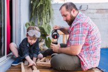 Hipster-Papa gießt Kräutertee aus Thermoskanne in Kalebassen-Kürbis gegen Jungen mit Bauholz auf Promenade — Stockfoto