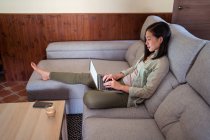 Vista lateral de la joven empleada remota étnica interesada trabajando en netbook en el sofá contra el café y el teléfono inteligente en casa - foto de stock