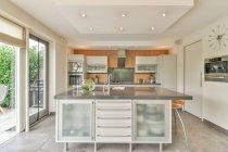 Интерьер современной кухни со столом и стульями против холодильника и шкафов в доме со стеклянной стеной в дневное время — стоковое фото