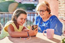 Щаслива бабуся сидить за столом і показує фотографії на смартфоні усміхненій бабусі, сидячи на терасі в сонячний день — стокове фото