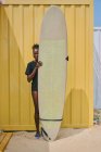 Allegro giovane sportiva afroamericana in bikini e t-shirt guardando la fotocamera con tavola da surf in un contenitore giallo sulla costa — Foto stock