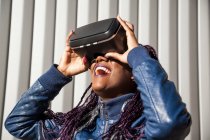 Задоволена молода афроамериканська жінка в VR хедсеті розважається і грає віртуальну гру проти сірої смугасті стіни — стокове фото