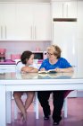 Concentrated grandmother in casual clothes and eyeglasses sitting at table and reading book with cheerful granddaughter in the kitchen at home — Stock Photo