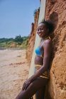 Seitenansicht einer afroamerikanischen Sportlerin, die mit einem Surfbrett von einem Strandabschnitt vor einem Lehmfelsen in die Kamera blickt — Stockfoto