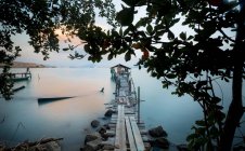 Blick durch Baumzweige auf einen Pier, der abends zu einer Hütte mitten auf dem Meer in Malaysia führt — Stockfoto