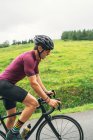 Вид сбоку спортсмена в защитном шлеме на велосипеде во время тренировки на асфальтовой дороге против зеленого холма и деревьев под легким небом — стоковое фото