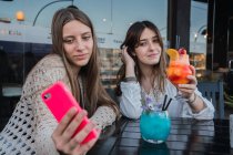 Кращі друзі-жінки в келихах освіжаючих напоїв беруть собі портрет на мобільний телефон за столом у міському кафе — стокове фото