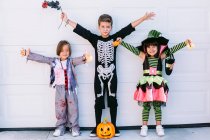 Fröhliche kleine Freunde in verschiedenen Halloween-Kostümen mit Kürbis und Accessoires heben die Arme und schauen in die Kamera, während sie an der weißen Wand zusammen stehen — Stockfoto