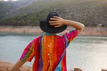 Rückansicht Frau blickt auf einen See mit einer Hand hält einen schwarzen Hut — Stockfoto