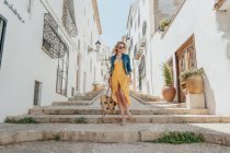 Full body front view of young female in summer outfit standing on stone stairway between old buildings in alley — Stock Photo