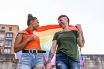Знизу веселі молоді гомосексуальні подруги з ЛГБТ-прапором, що тримаються за руки, дивлячись один на одного і крокуючи по міському тротуару. — стокове фото