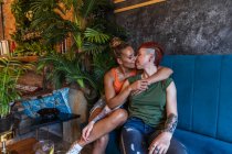 Zufriedene junge tätowierte Frau mit Mohawk und Getränk umarmt und küsst lesbische Freundin, während sie sich auf der Couch im Haus anschaut — Stockfoto