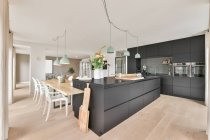 Стильный интерьер просторной кухни с черной мебелью и деревянным столом в современной квартире в дневное время — стоковое фото
