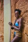 Vue latérale d'une athlète afro-américaine regardant une caméra avec une planche de surf depuis une zone de la plage et devant un rocher argileux — Photo de stock