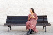 Full body of positive female in stylish clothes browsing smartphone and sitting on wooden bench on street in daytime — Stock Photo