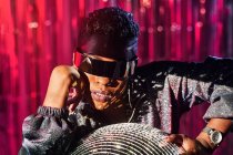 Femme ethnique tendance en lunettes cyberpunk et montre-bracelet touchant boule disco en rayon clair — Photo de stock