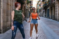 Side view of happy young homosexual female couple with tattoos in trendy clothes looking at each other while having fun on street — Stock Photo