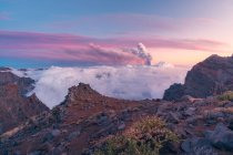 La salida del sol en los picos poderosos de la montaña en medio de nubes blancas gruesas suaves y en el fondo la erupción de un volcán. Cumbre Vieja erupción volcánica en La Palma Islas Canarias, España, 2021 - foto de stock