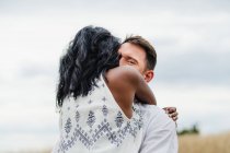 Hombre feliz abrazando novia india de pie en el campo bajo el cielo nublado - foto de stock