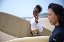 Sonriente deportista negra con longboard contra novia asiática con tabla de surf mirando hacia adelante en el océano bajo el cielo azul nublado - foto de stock