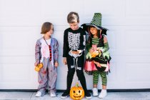 Full body of group of little kids dressed in various Halloween costumes with carved Jack O Lantern browsing mobile phone together while standing near white wall on street — Stock Photo
