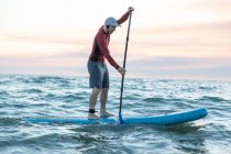 Vue latérale du surfeur masculin en combinaison et chapeau sur planche à pagaie surfant sur le bord de mer pendant le coucher du soleil — Photo de stock
