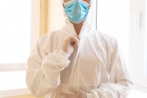 Cropped unrecognizable male medic in personal protective equipment with glasses and sterile mask looking forward against window in hospital — Stock Photo