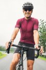 Веселый взрослый спортсмен в велосипедных солнцезащитных очках и шлеме, сидящий на дорожном велосипеде на сельской дороге при дневном свете — стоковое фото