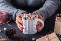 Dall'alto di raccolto femmina che tiene la tazza di bevanda calda con marshmallows tra coni di regali di Natale e bastoni di cannella — Foto stock