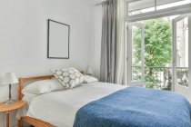 Interior do quarto elegante com cama confortável perto da janela e paredes claras durante o dia — Fotografia de Stock