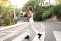 Seitenansicht einer freundlichen jungen Frau in Gläsern mit blühendem Blumenstrauß, die wegschaut, wenn sie die Fahrbahn überquert — Stockfoto