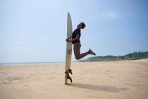 Vista lateral de la mujer surfista afroamericana feliz en tanga con longboard surf saltar por encima de la costa arenosa bajo el cielo azul nublado - foto de stock