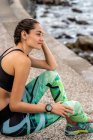 Vue latérale d'une athlète féminine mince en tenue de sport assise sur une promenade en pierre près de la mer et faisant une pause pendant l'entraînement — Photo de stock