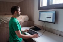 Vista lateral do blogueiro masculino em óculos edição de texto no monitor enquanto digita no teclado na sala da casa — Fotografia de Stock