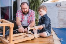 Бородатый отец учит сына молотком работать с деревом, сидя на набережной в выходные — стоковое фото
