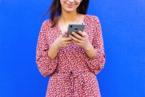 Обрізана невпізнавана усміхнена жінка в сукні та окулярах, що стоїть біля синьої стіни та використовує смартфон удень — стокове фото