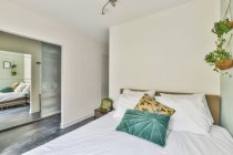 Dormitorio contemporáneo con almohadas en edredón entre plantas en maceta en casa con suelo de baldosas - foto de stock