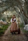 Серьёзная ведьма в платье стоит с волшебной книгой заклинаний и метлой на дороге в лесу и смотрит в сторону — стоковое фото