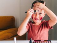 Charming child with makeup applicator touching head while looking at camera at table with eyeshadow palette — Stock Photo