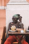 Hombre afroamericano maduro fresco en gafas de sol y pulseras sentados en la mesa de la cafetería urbana con bebida de coco - foto de stock