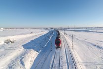Drone view of train riding on railroad through snowy terrain under blue clear sky — Stock Photo