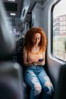 Femme intéressée avec les cheveux bouclés en jeans déchiré messagerie texte sur téléphone portable pendant le voyage en train en journée — Photo de stock