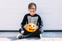 Sorrindo menino pré-adolescente vestindo traje de Halloween preto com esqueleto impressão de pé perto esculpida Jack O Lanterna abóbora contra a parede branca — Fotografia de Stock