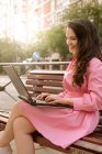 Giovane donna d'affari allegra in abito seduto sulla panchina a lavorare su netbook sulla strada della città — Foto stock