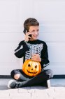 Full body of cheerful boy in black skeleton costume with painted face and carved Halloween pumpkin talking on mobile phone while sitting near white wall on street — Stock Photo