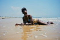 Весела молода етнічна жінка в купальнику з афро волоссям, дивлячись далеко, лежачи на узбережжі океану під блакитним небом — стокове фото