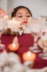 Niño tierno contemplando vela encendida en vidrio en la mesa con conos de coníferas durante las vacaciones de Año Nuevo en casa - foto de stock